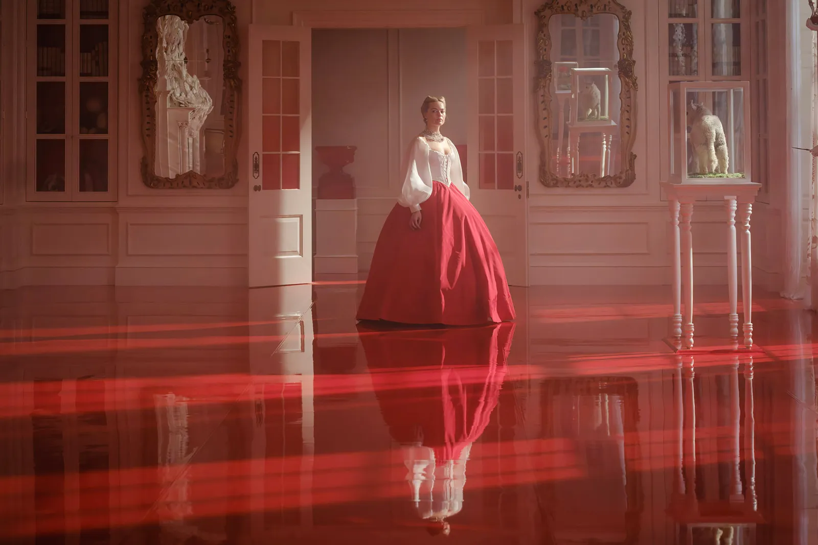 Margot Robbie as Catherine Earnshaw standing in a grand red-lit interior wearing a white corset and dramatic red skirt — directed by Emerald Fennell, from Wuthering Heights (2026), based on the novel by Emily Brontë.