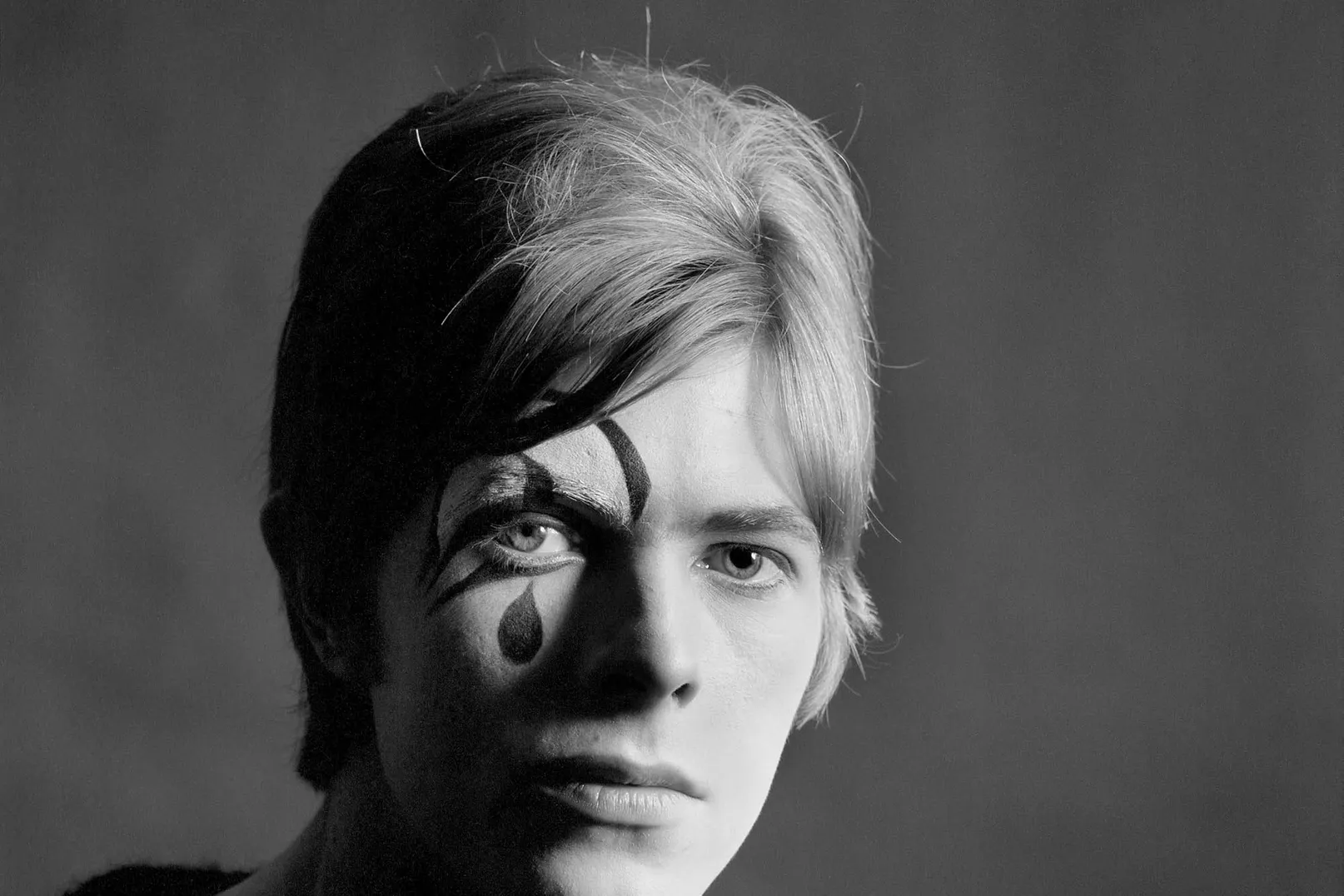 Black-and-white studio portrait of David Bowie as a young performer, looking directly at the camera with short light hair and theatrical face paint—a bold curved stripe above one eye and a dark teardrop below it. Photo: Gerald Fearnley/Gerald Fearnley.