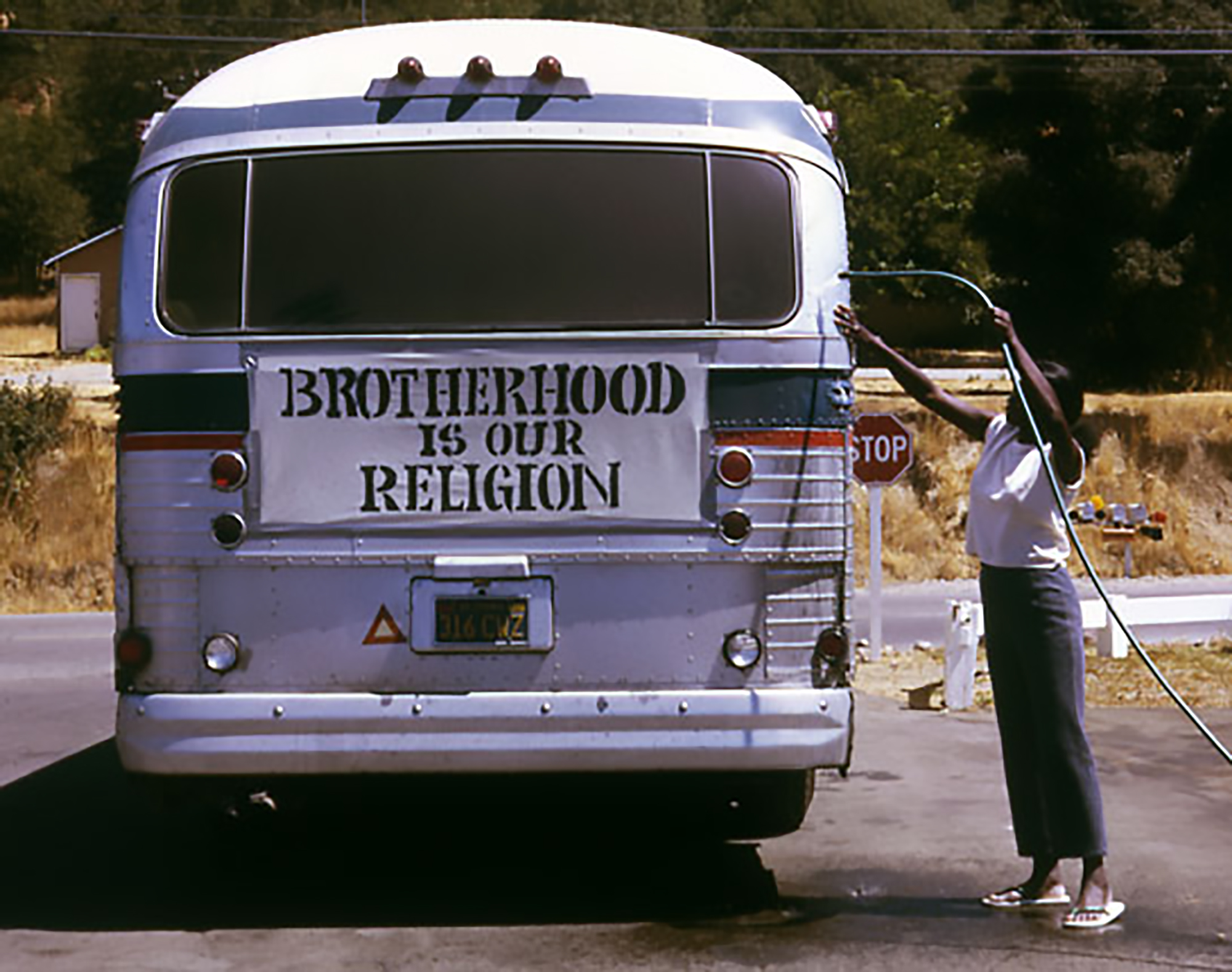 A Peoples Temple bus at a roadside stop, displaying a large banner that reads “Brotherhood Is Our Religion,” with a woman standing beside the vehicle.